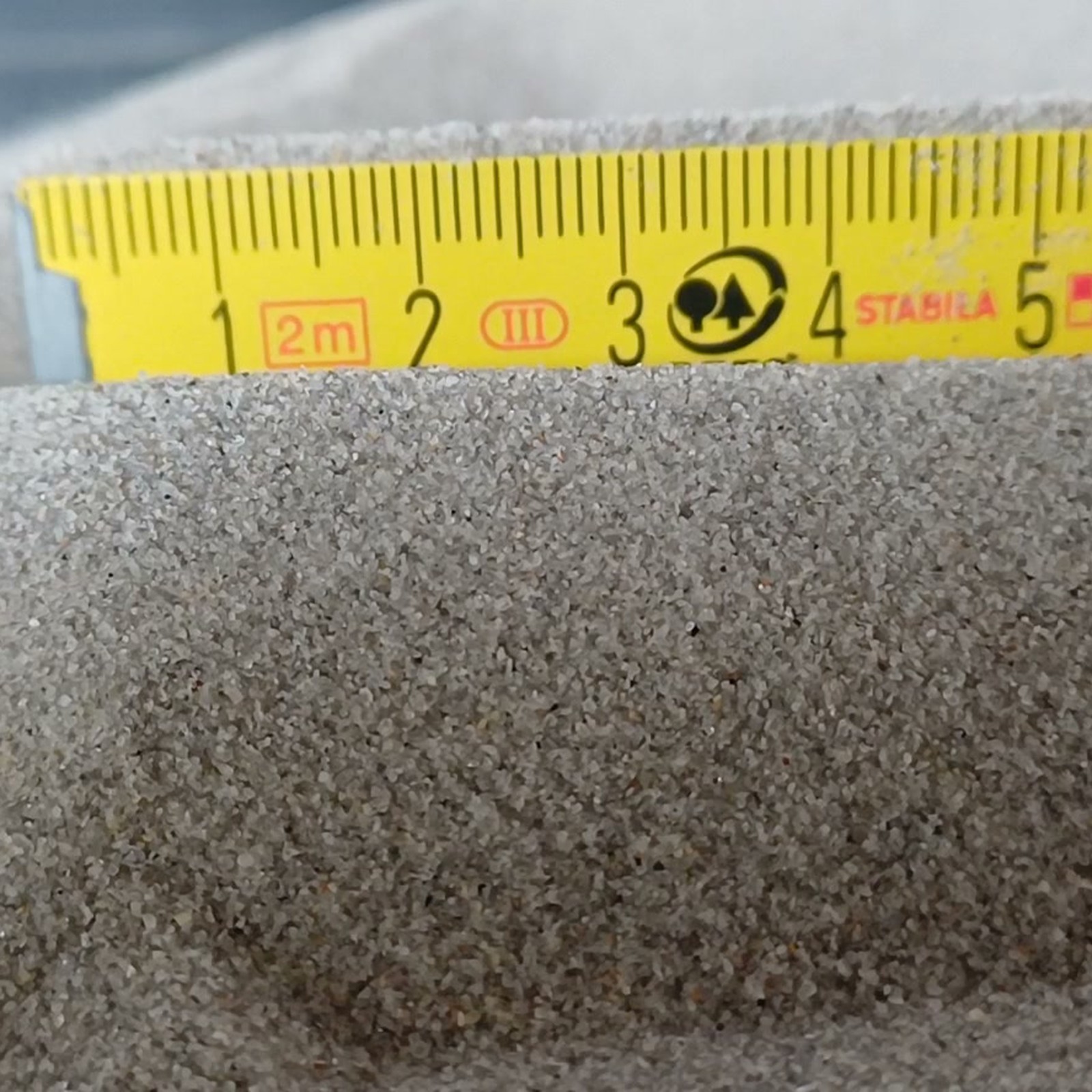 Chinchillasand Naturgrau, 0,1–0,3 mm, 5 kg: Grauer Sandrücken mit Maßstab dahinter.