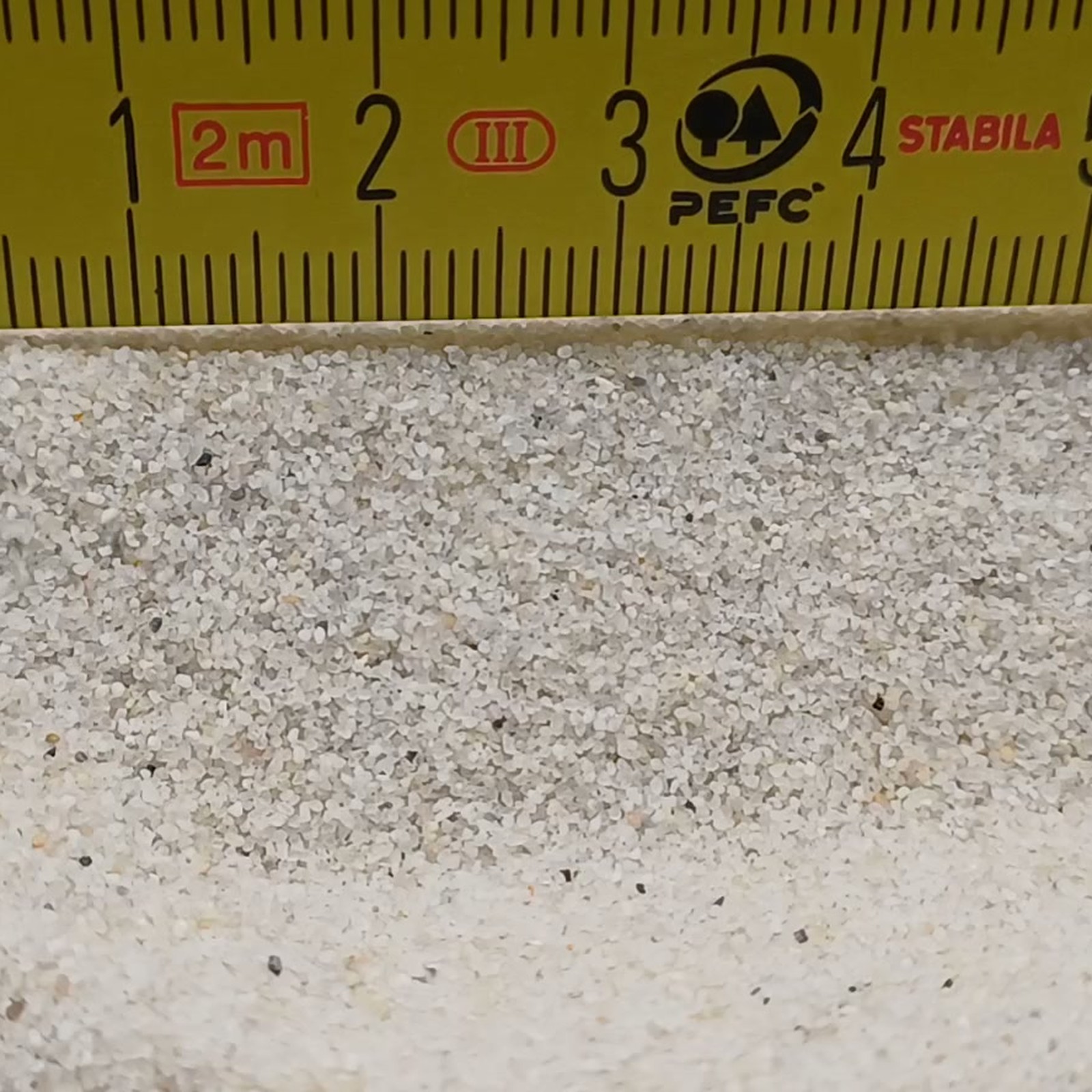 Aquariumsand Naturweiß, 0,2–0,5 mm, 25 kg: Helle Kornfläche vor gelbem Gliedermaßstab.