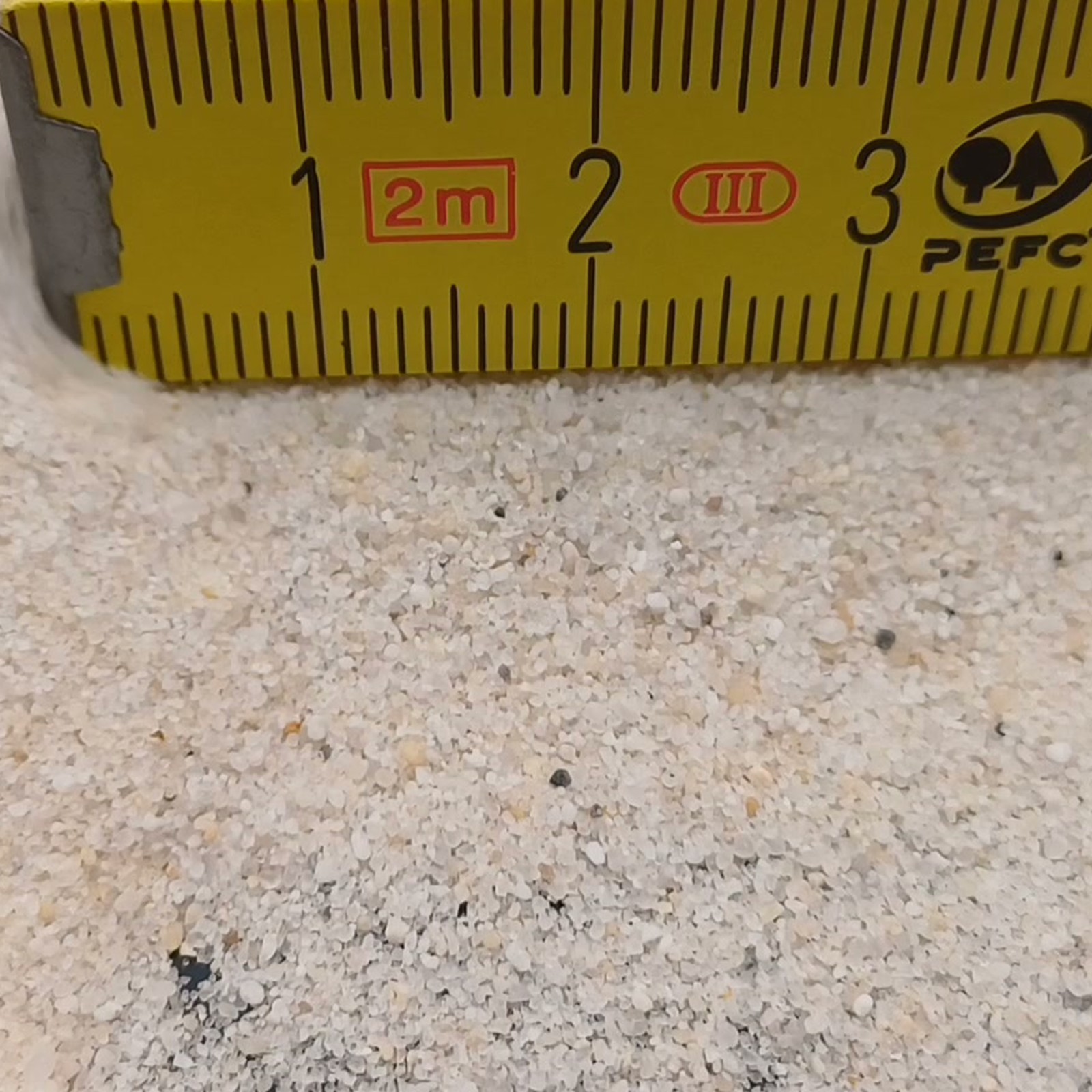 Industriesand Naturweiß, 0,1–0,6 mm, 25 kg: Sandkörner auf dunklem Untergrund vor dem Maßstab.