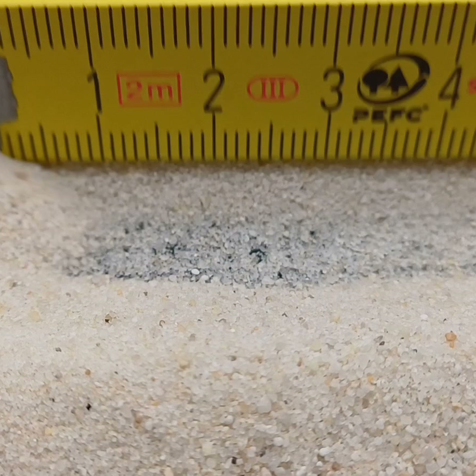 Industriesand Naturweiß, 0,1–0,6 mm, 25 kg: Flache Sandrinne unter einem gelben Maßstab.