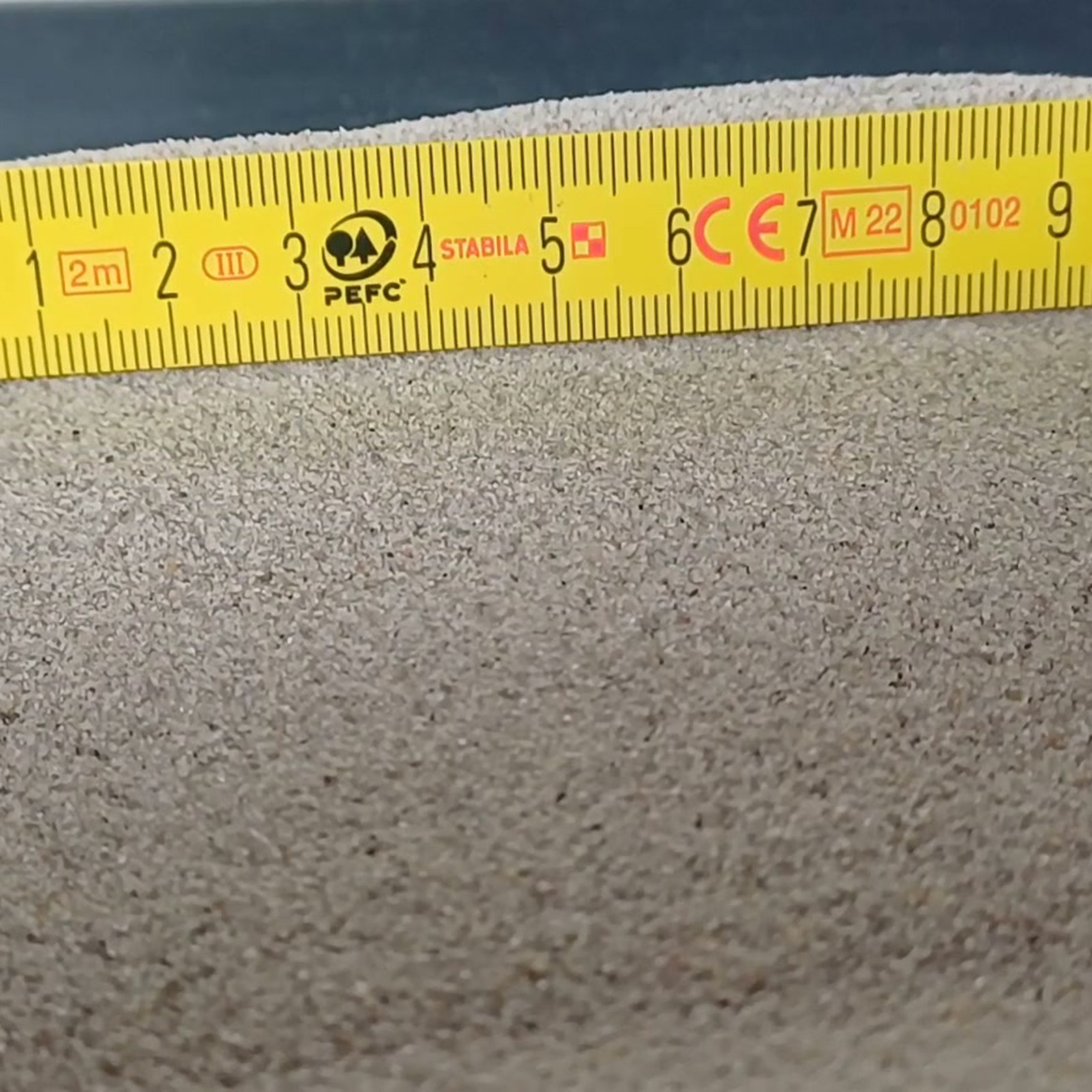 Aquariumsand Naturgrau, 0,1–0,5 mm, 10 kg: Graue Sandfläche vor einem gelben Gliedermaßstab.