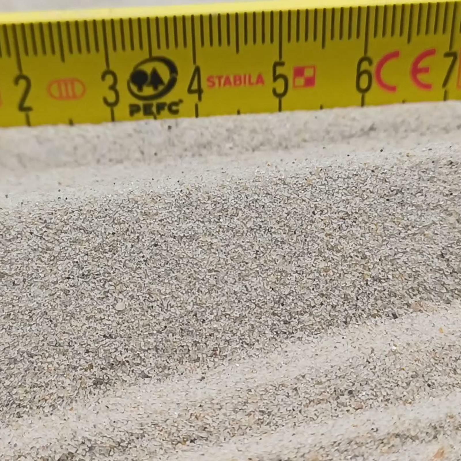 Chinchillasand Naturbeige, 0,1–0,3 mm, 25 kg: Sandkante vor einem gelben Maßstab.