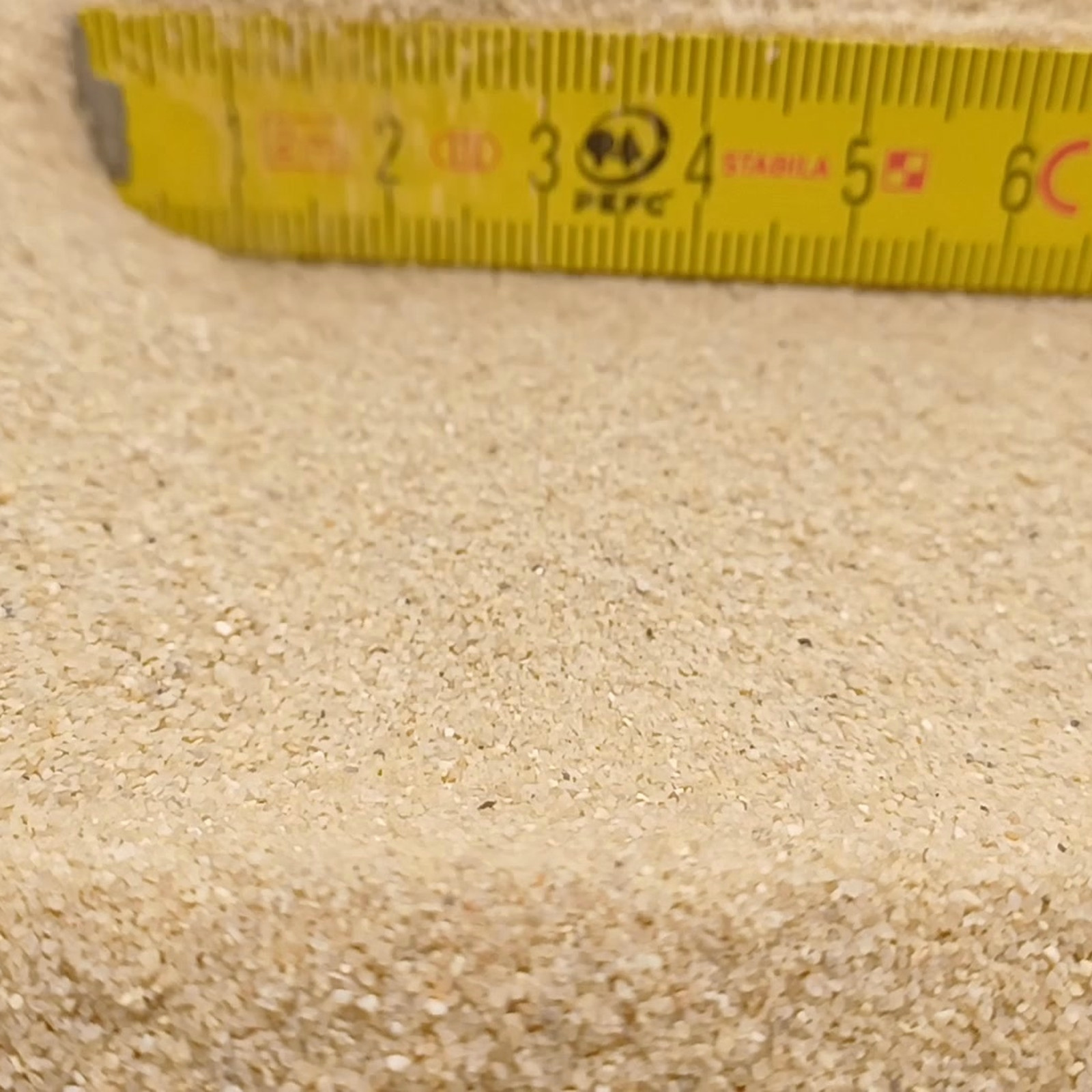 Aquariumsand Naturbeige, 0,1–0,6 mm, 25 kg: Feine Kornfläche vor einem gelben Gliedermaßstab.