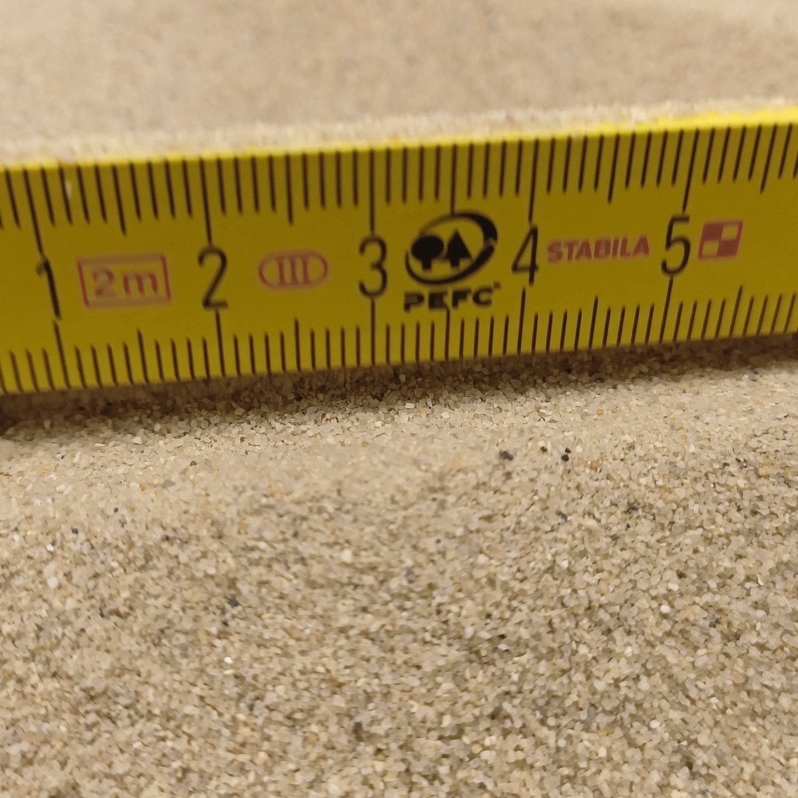 Terrariumsand Naturbeige, 0,3–0,6 mm, 25 kg: Feine Sandkörner vor einem gelben Gliedermaßstab.